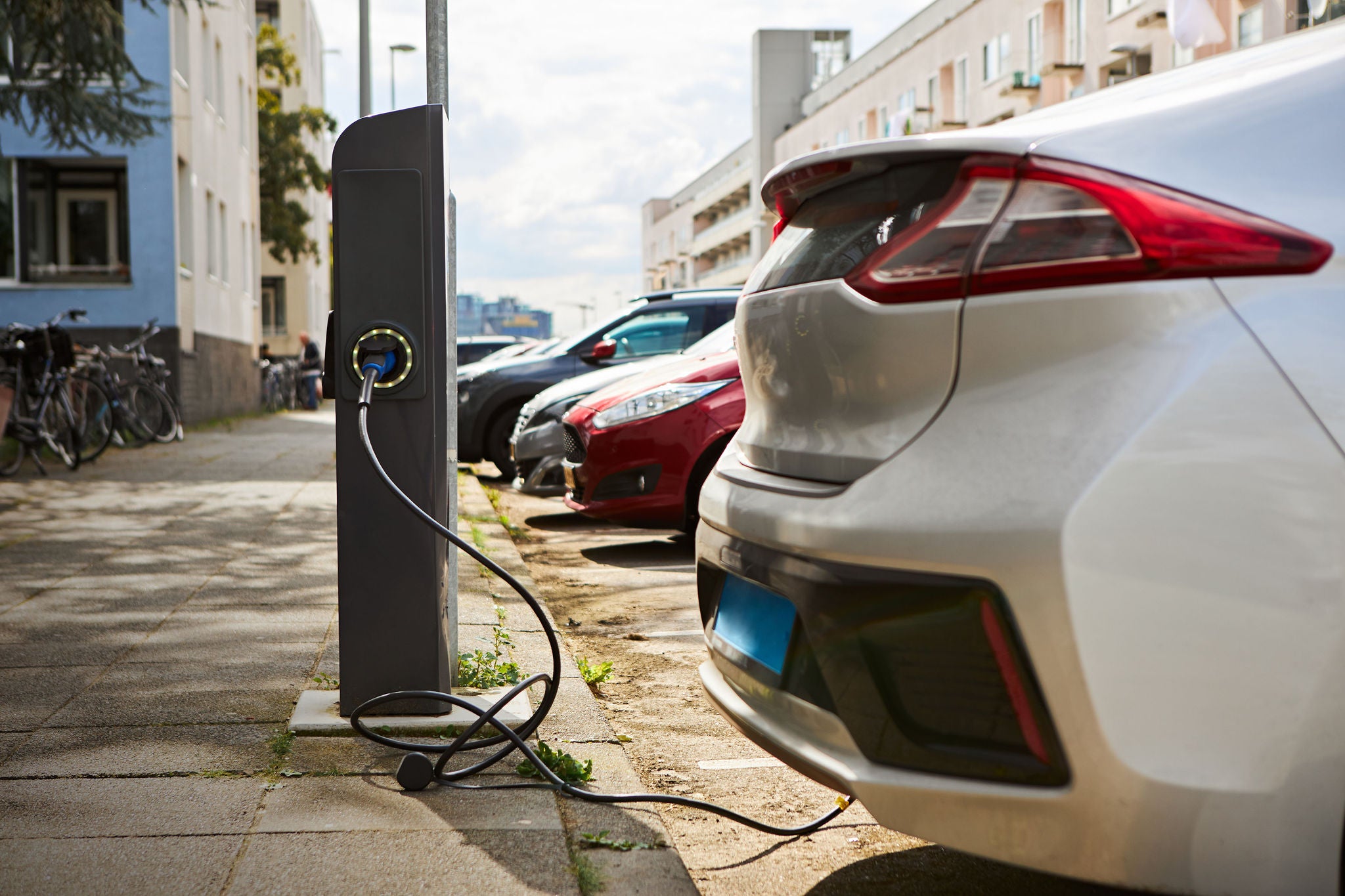 Cropped shot of silver electric car at charging station with power cable supply plugged in against cityscape background. EV fuel, automobile industry, modern technology and clean energy concept