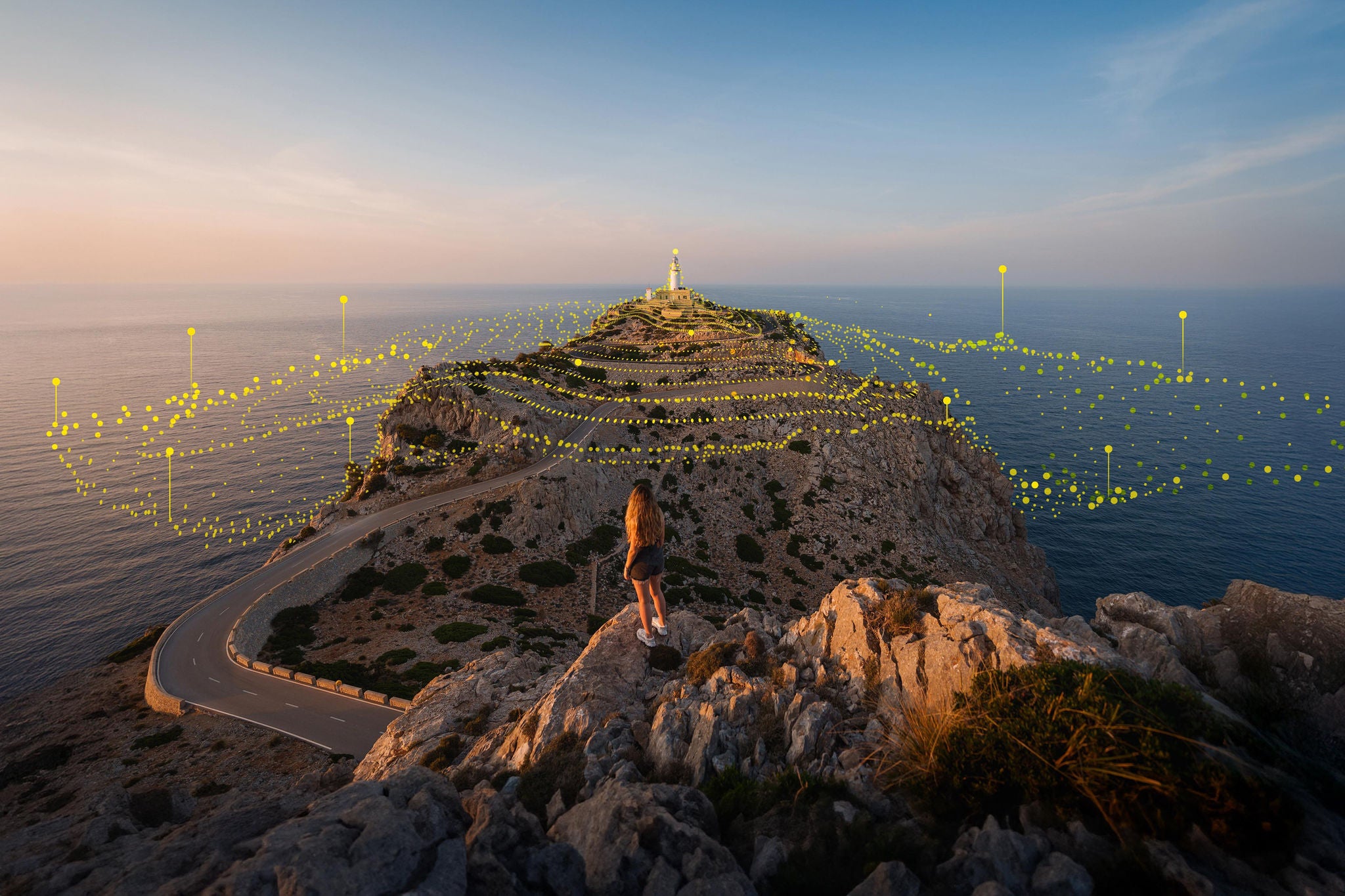 Young woman standing on a cliff looking at cabo formentor lighthouse at sunset in mallorca, balearic islands, spain