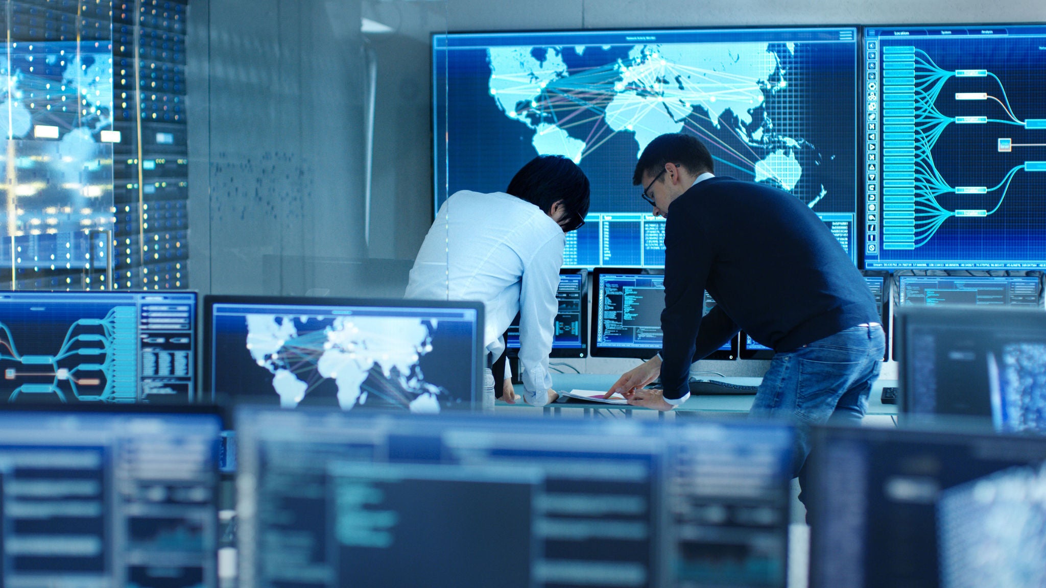 Na sala de controle do sistema, o gerente de projeto e o engenheiro de TI conversam, cercados por vários monitores com gráficos. O Big Monitor mostra um mapa interativo de logística.